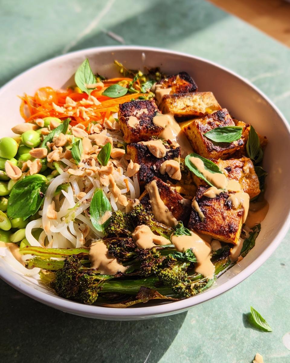 A vibrant bowl featuring crispy tofu, roasted broccoli, edamame, shredded carrots, and noodles, drizzled with a creamy sauce, part of 20-Ingredient Rice Bowls Recipes.