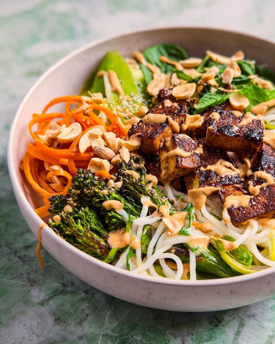 A vibrant bowl of 20-ingredient rice bowl featuring crispy tofu, broccolini, shredded carrots, and avocado with a drizzle of peanut sauce.