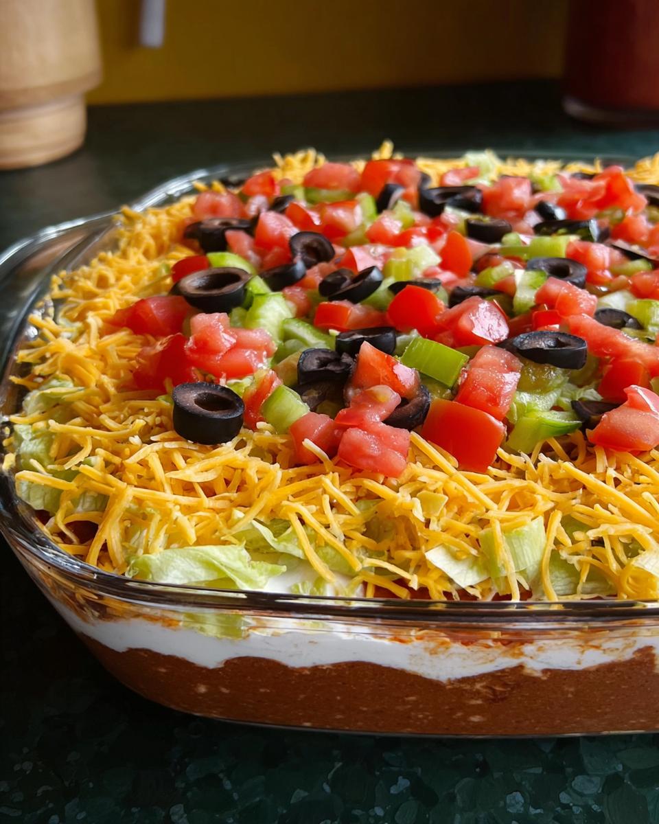 A close-up of a 7-layer taco dip, a popular Taco Tuesday recipe, featuring layers of beans, sour cream, cheese, lettuce, tomatoes, olives, and green peppers.