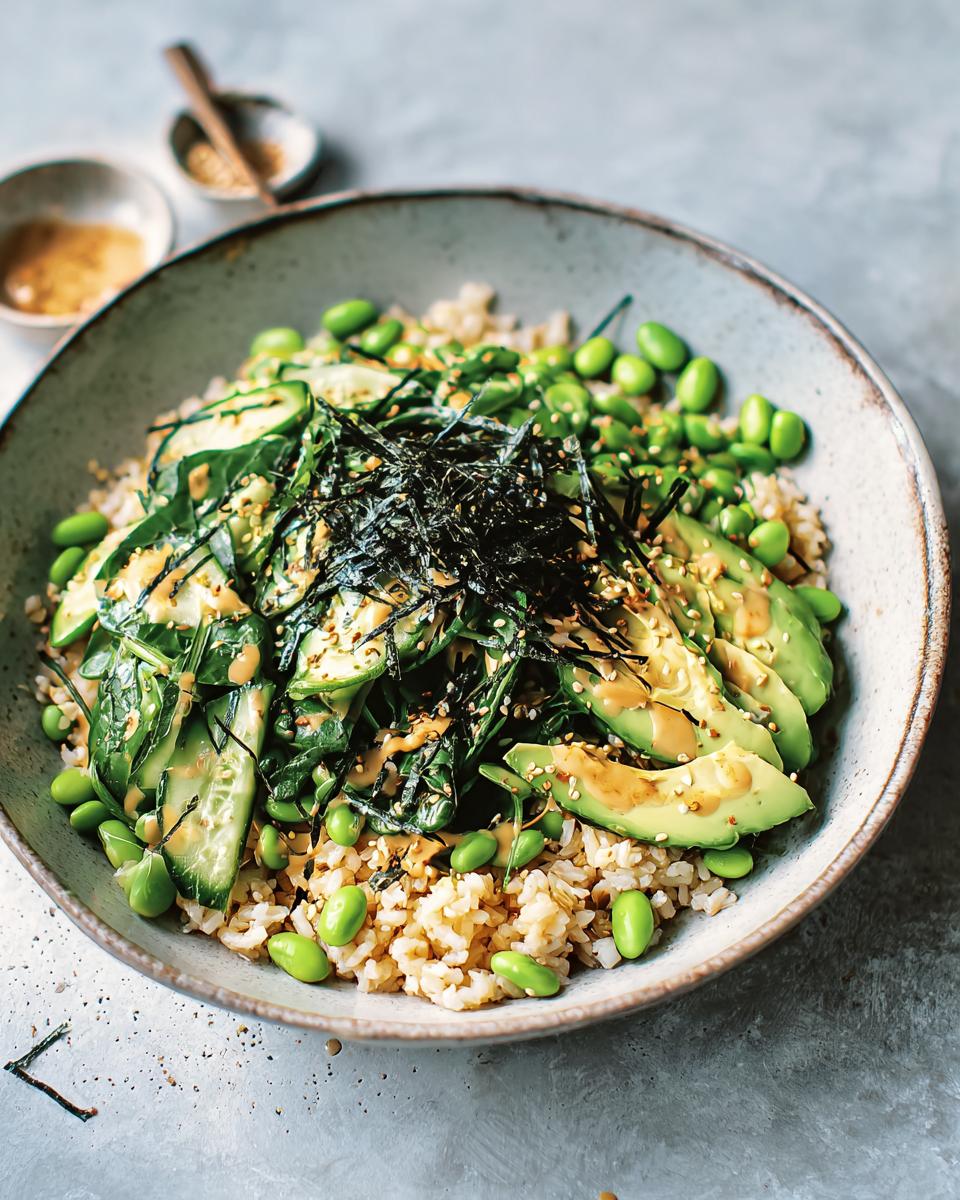 A vibrant rice bowl featuring brown rice, sliced avocado, edamame, cucumber, and nori, drizzled with a creamy sauce.