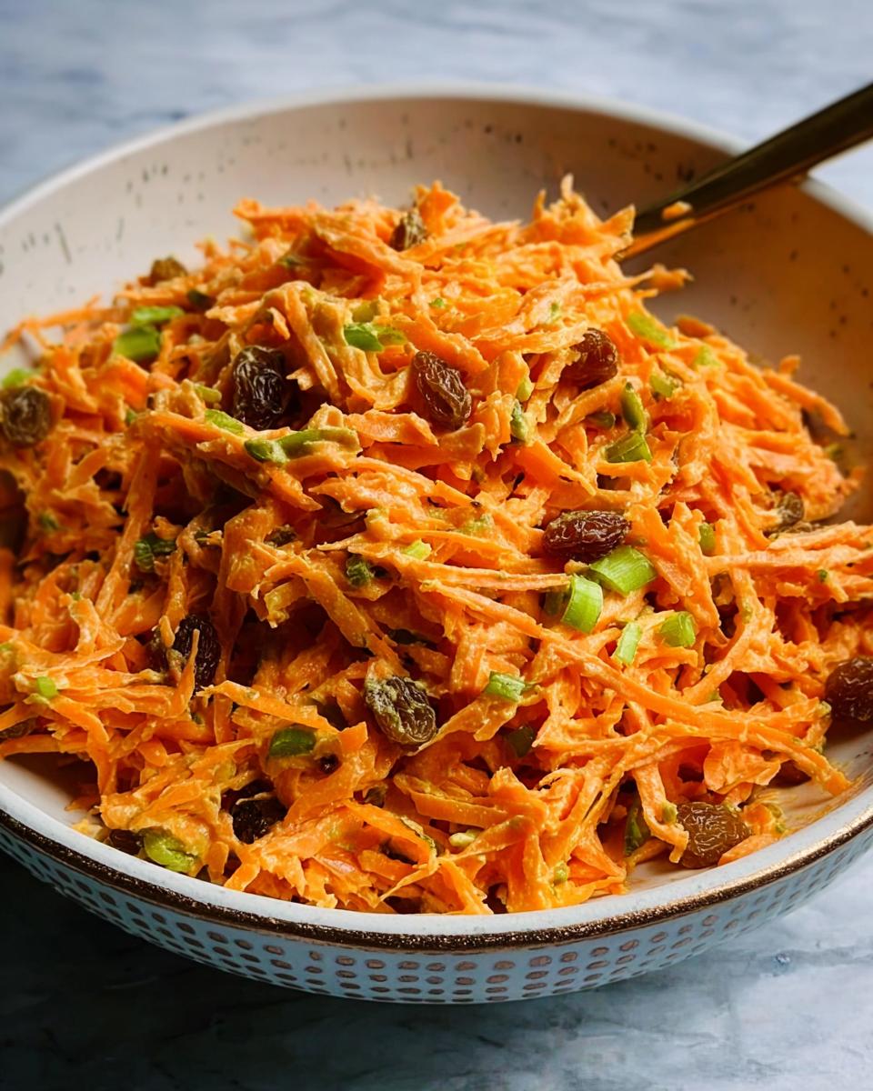 Close-up of a bowl filled with shredded carrot salad, mixed with raisins and chopped green onions. A quick veggie side.