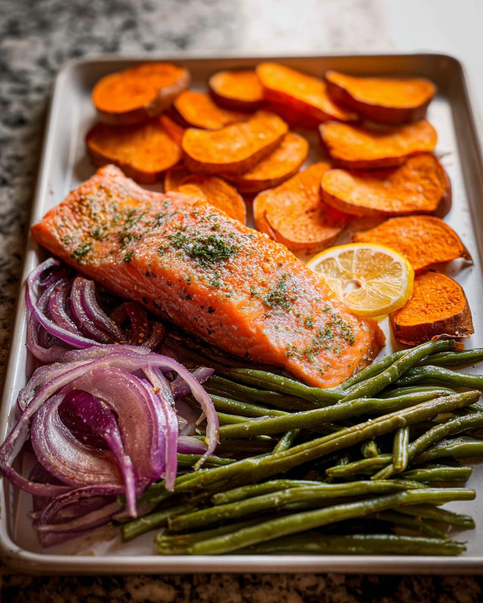 A perfectly cooked salmon fillet on a baking sheet with roasted sweet potato slices, green beans, red onion, and a lemon wedge.