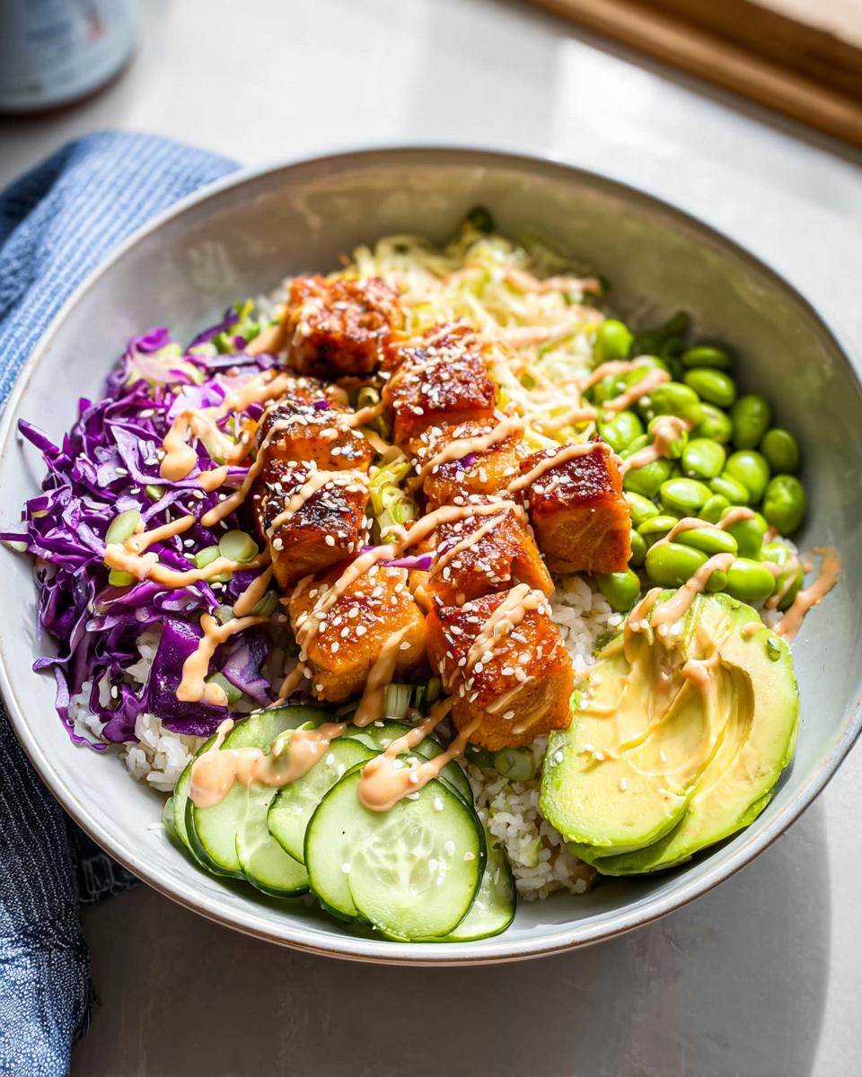 A vibrant Crispy Salmon and Rice Bowl featuring glazed salmon cubes, rice, edamame, red cabbage, cucumber, and avocado, drizzled with sauce.