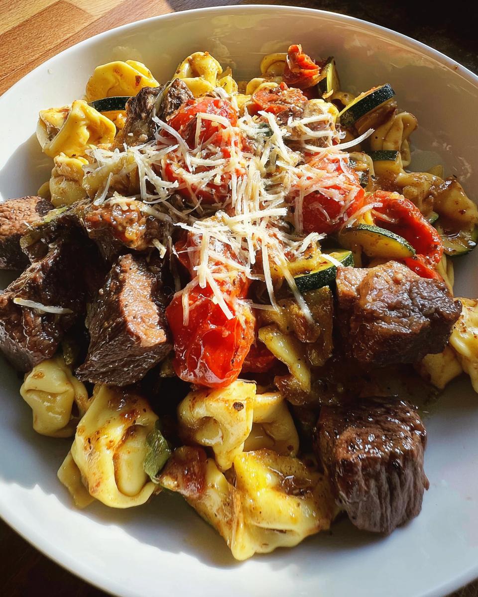 A close-up of a bowl of Garlic Steak Tortellini, featuring tender steak pieces, tortellini pasta, cherry tomatoes, and zucchini, topped with grated cheese.