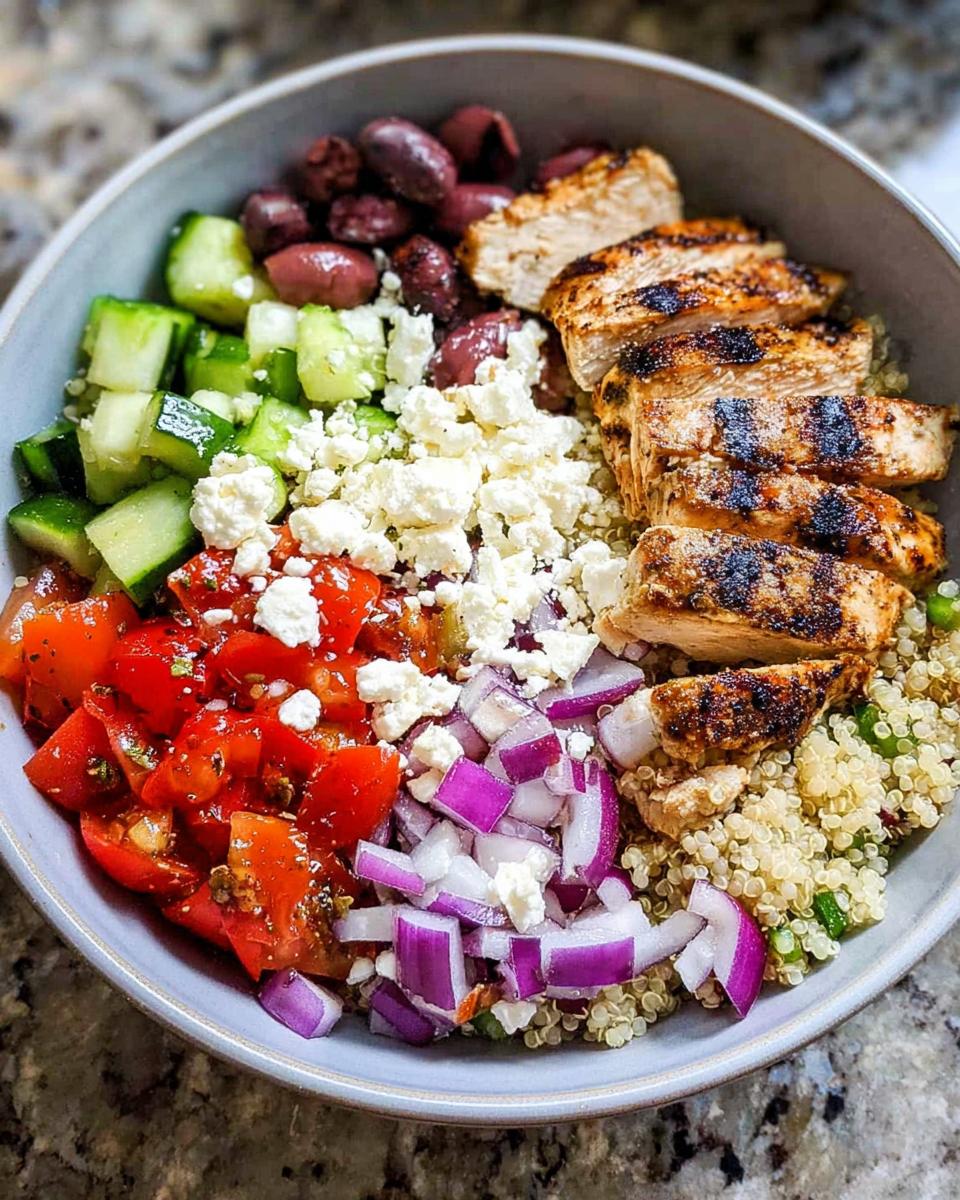 A close-up of a Greek Chicken Bowl with grilled chicken, quinoa, tomatoes, cucumber, olives, feta, and red onion.