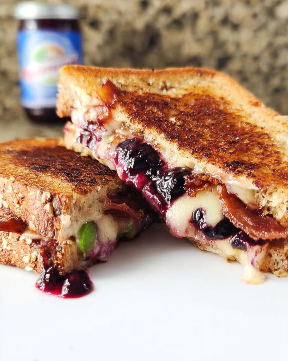 A gourmet blueberry grilled cheese sandwich cut in half, revealing melted cheese, blueberries, and bacon. Drips of blueberry jam are visible.