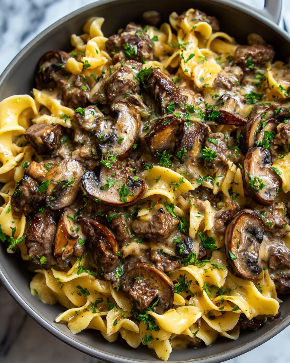 Close-up of a creamy beef stroganoff skillet meal with wide egg noodles, tender beef chunks, sautéed mushrooms, and fresh parsley.