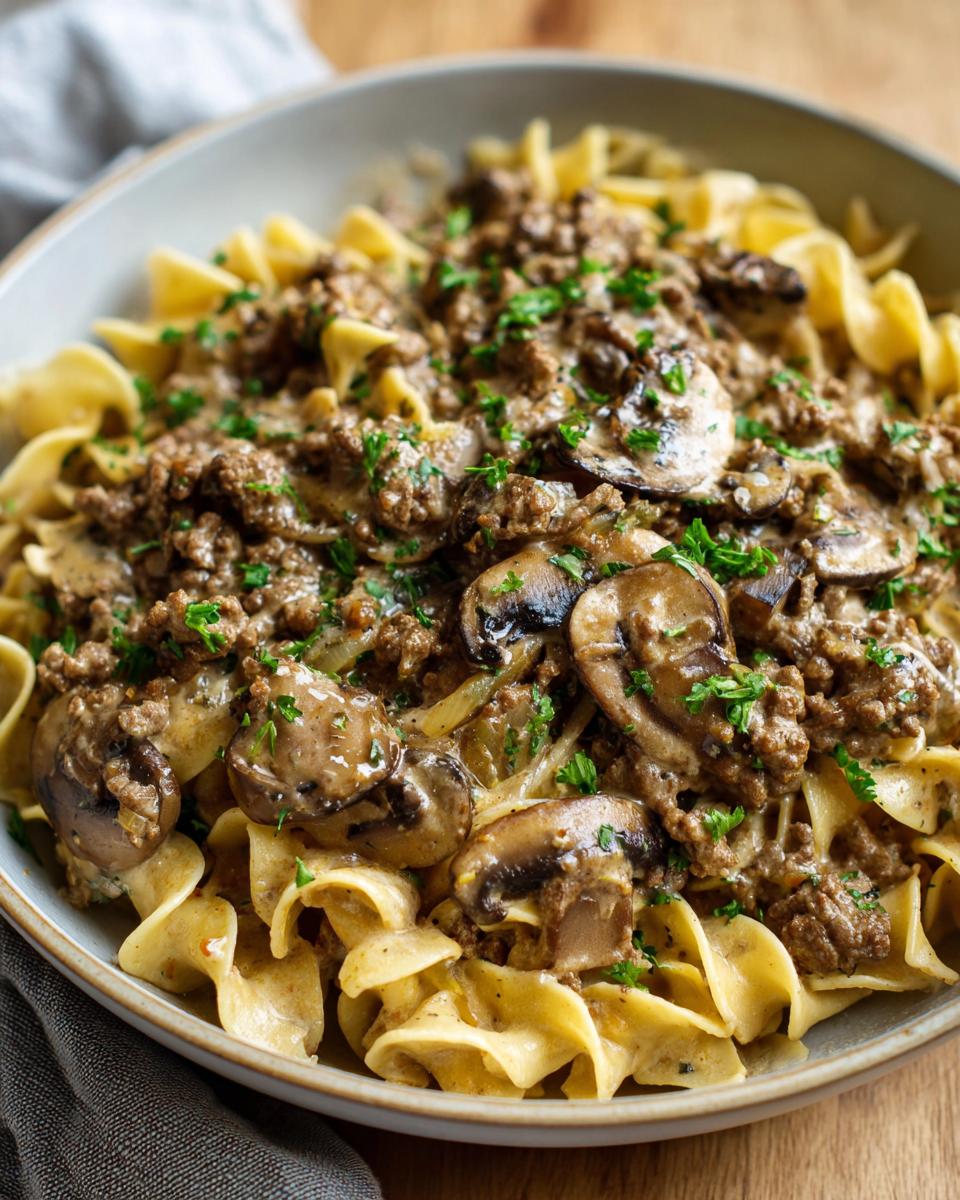 A close-up of a bowl of creamy beef stroganoff with ground beef, mushrooms, and egg noodles, garnished with parsley.