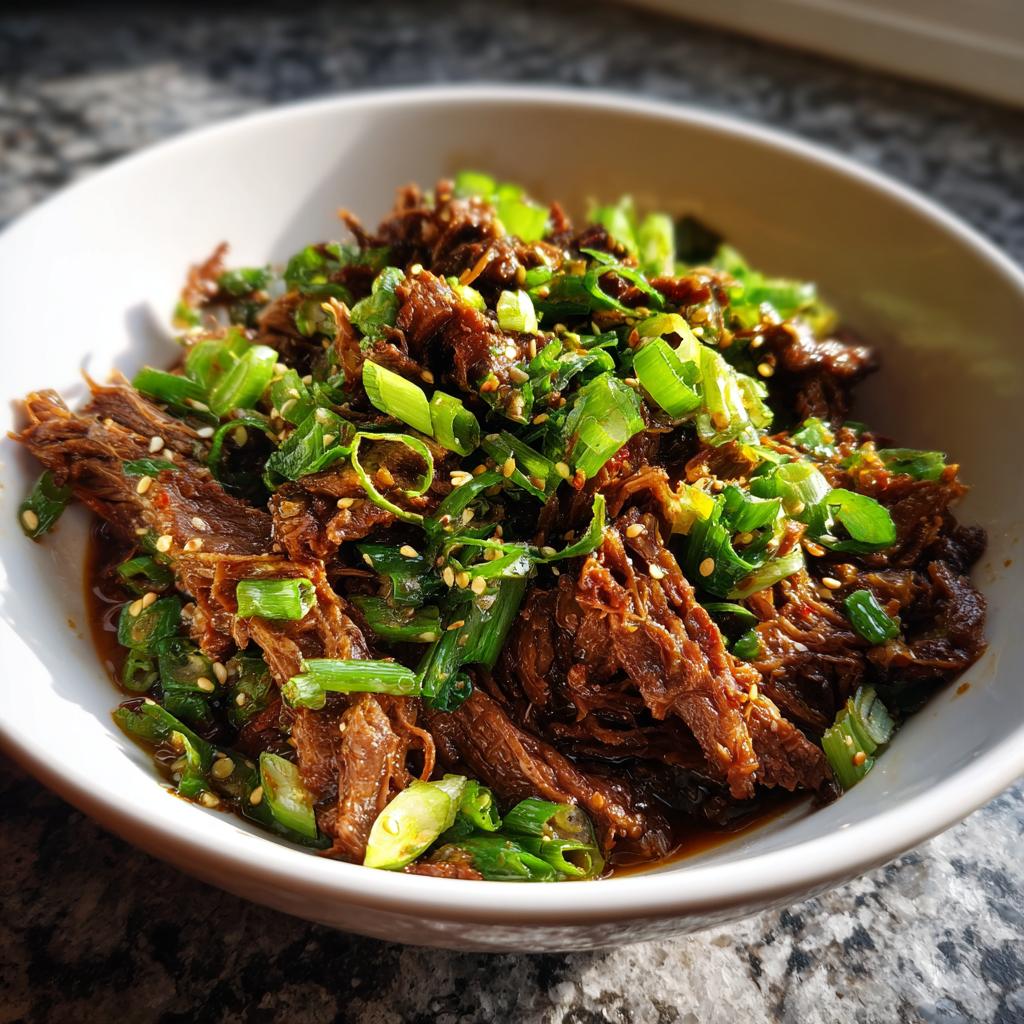A close-up of shredded Korean Style Pot Roast in a white bowl, topped with chopped scallions and sesame seeds.