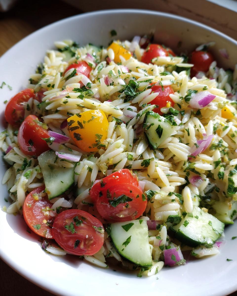 A close-up of a white bowl filled with lemony orzo salad, featuring cherry tomatoes, cucumber, red onion, and parsley.